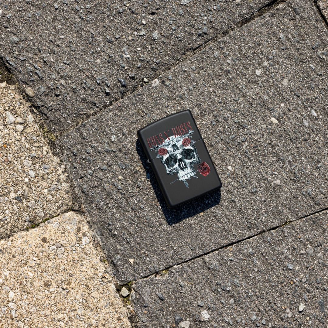 Lifestyle image of Zippo Guns N' Roses Flower Skull Design Black Matte Windproof Lighter lying on pavestone showing the front of the lighter. 

