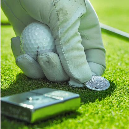 Lifestyle image of Zippo Golf Ball Marker Emblem – Zippo Design Street Chrome Windproof Lighter lying flat on the golf course showing the front, bottom stamp and non-hinge side of the lighter while the golf ball marker is being placed on the green as a ball marker. 
