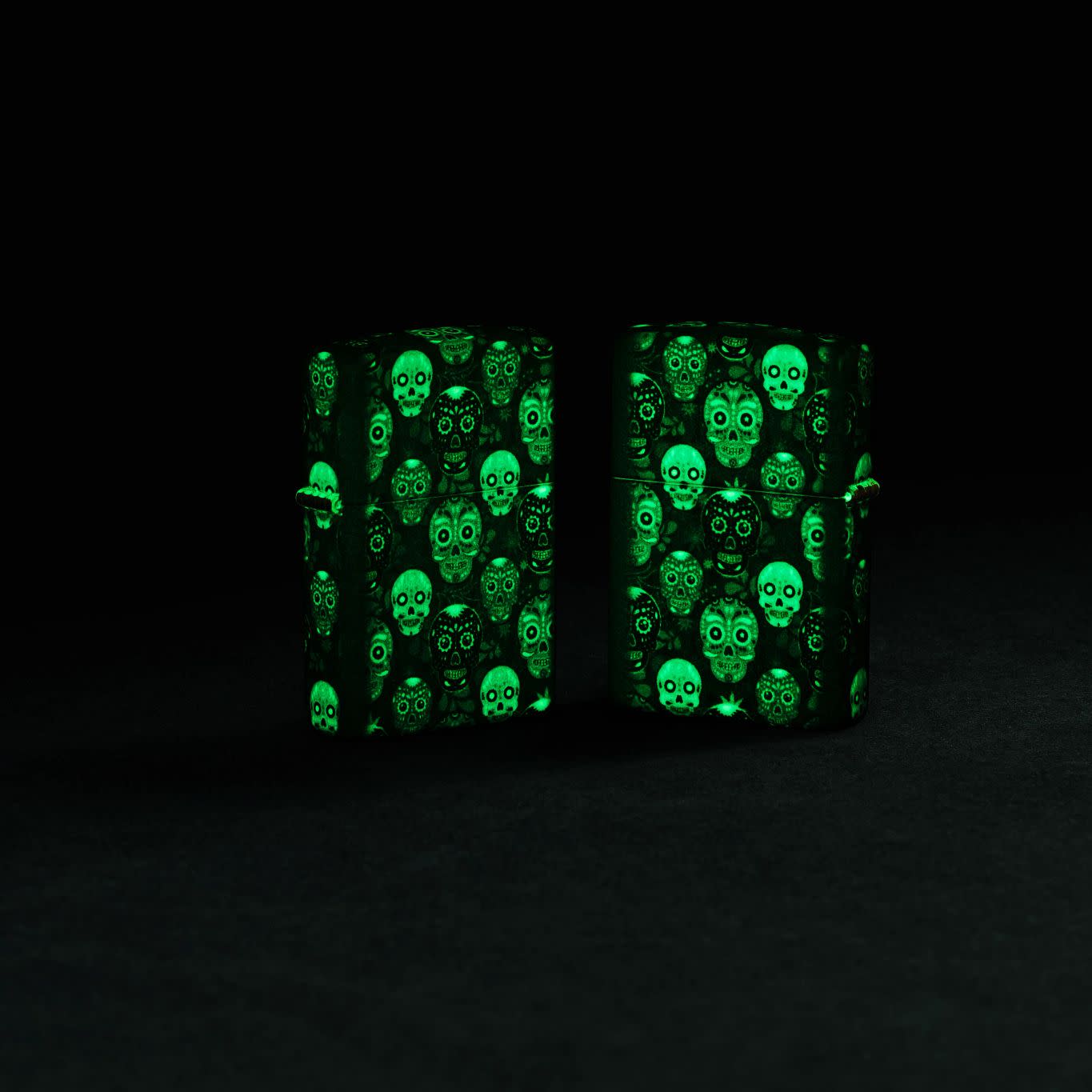 Lifestyle image of two Zippo Sugar Skulls Design Glow in the Dark Matte Windproof Lighters glowing in the dark.