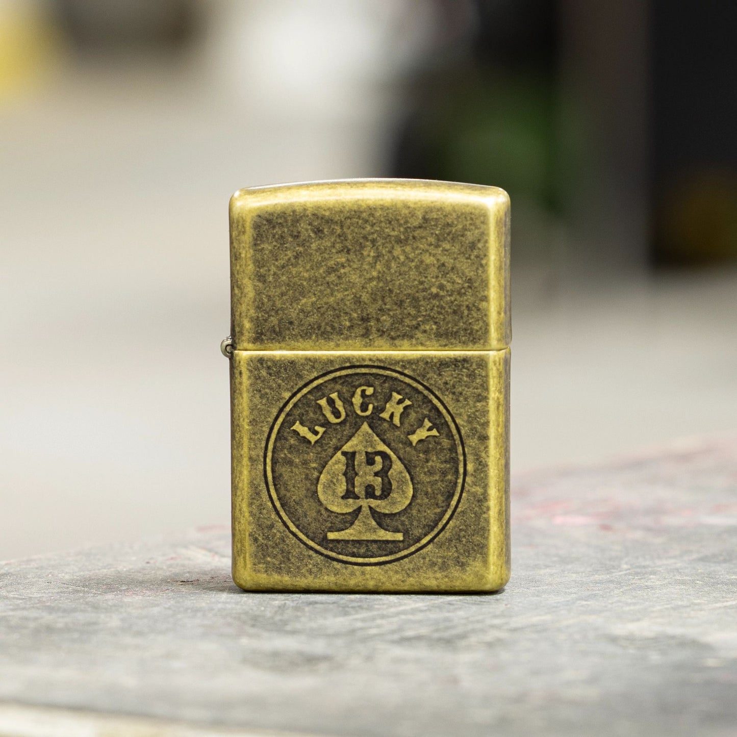 Lifestyle image of Zippo Lucky 13 Design Antique Brass Windproof Lighter standing on a concrete surface showing the front of the lighter. 

