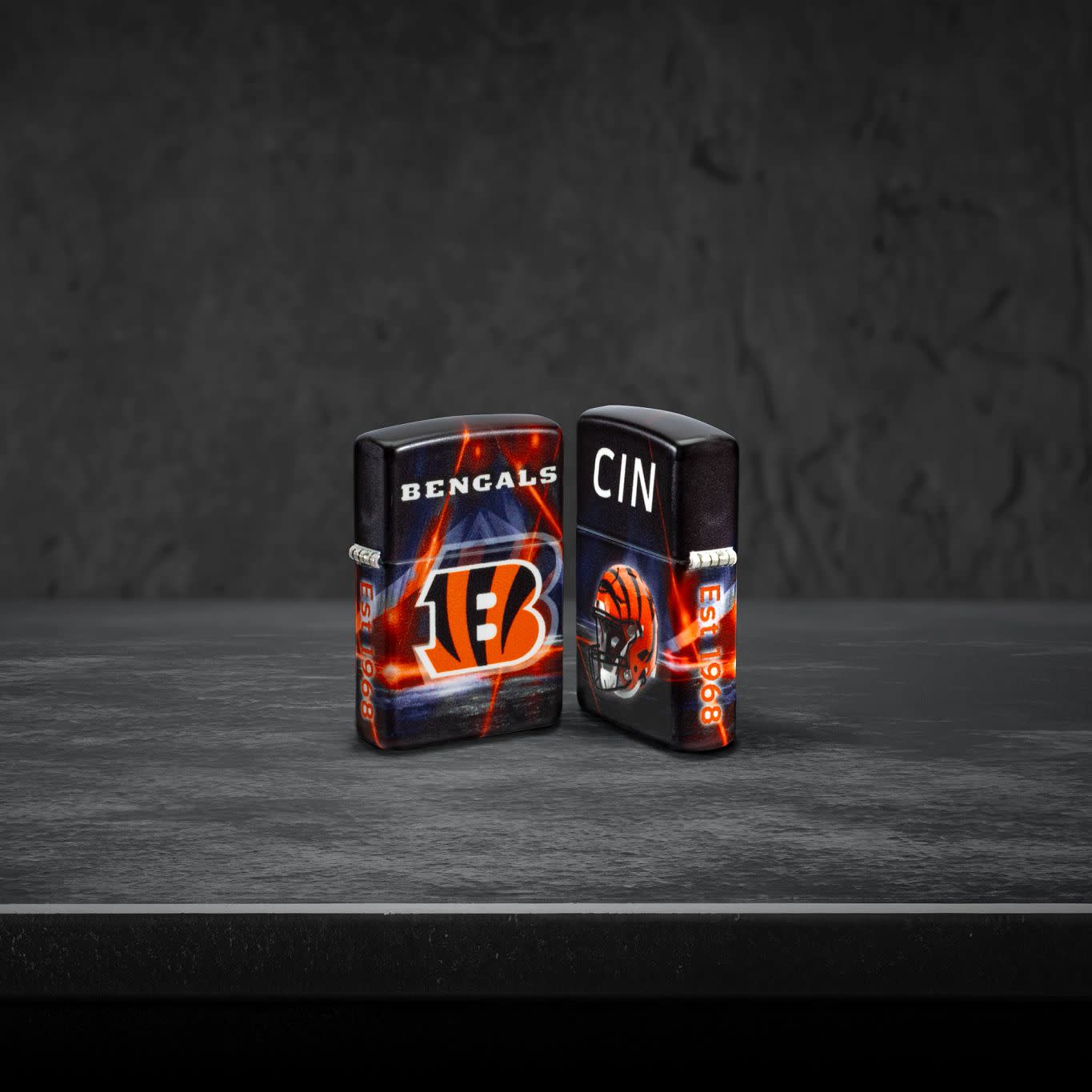 Lifestyle image of two Zippo NFL Cincinnati Bengals 540 Matte Windproof Lighters, one showing the front of the lighter and the other showing the back, standing on a dark grey surface.