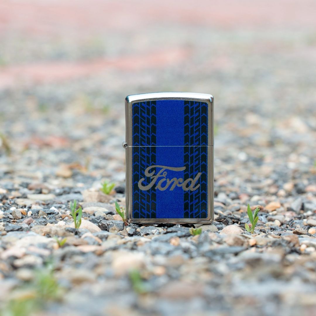 Lifestyle image of Zippo Ford Racing Stripes Design Street Chrome Windproof Lighter standing on gravel showing the front of the lighter with an out of focus background. 

