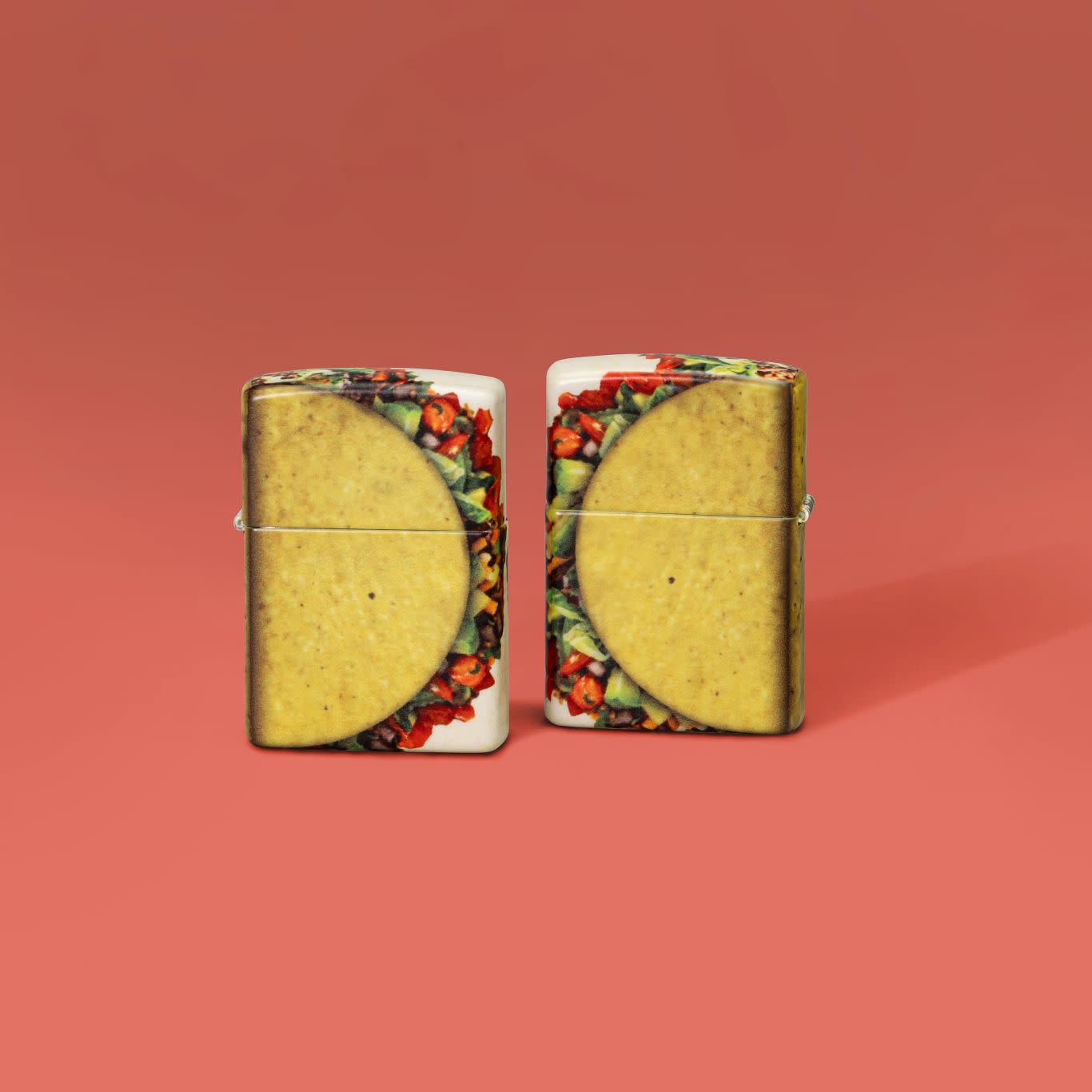 Lifestyle image of two Zippo Loaded Taco Design 540 Matte Windproof Lighters set on a reddish orange background.