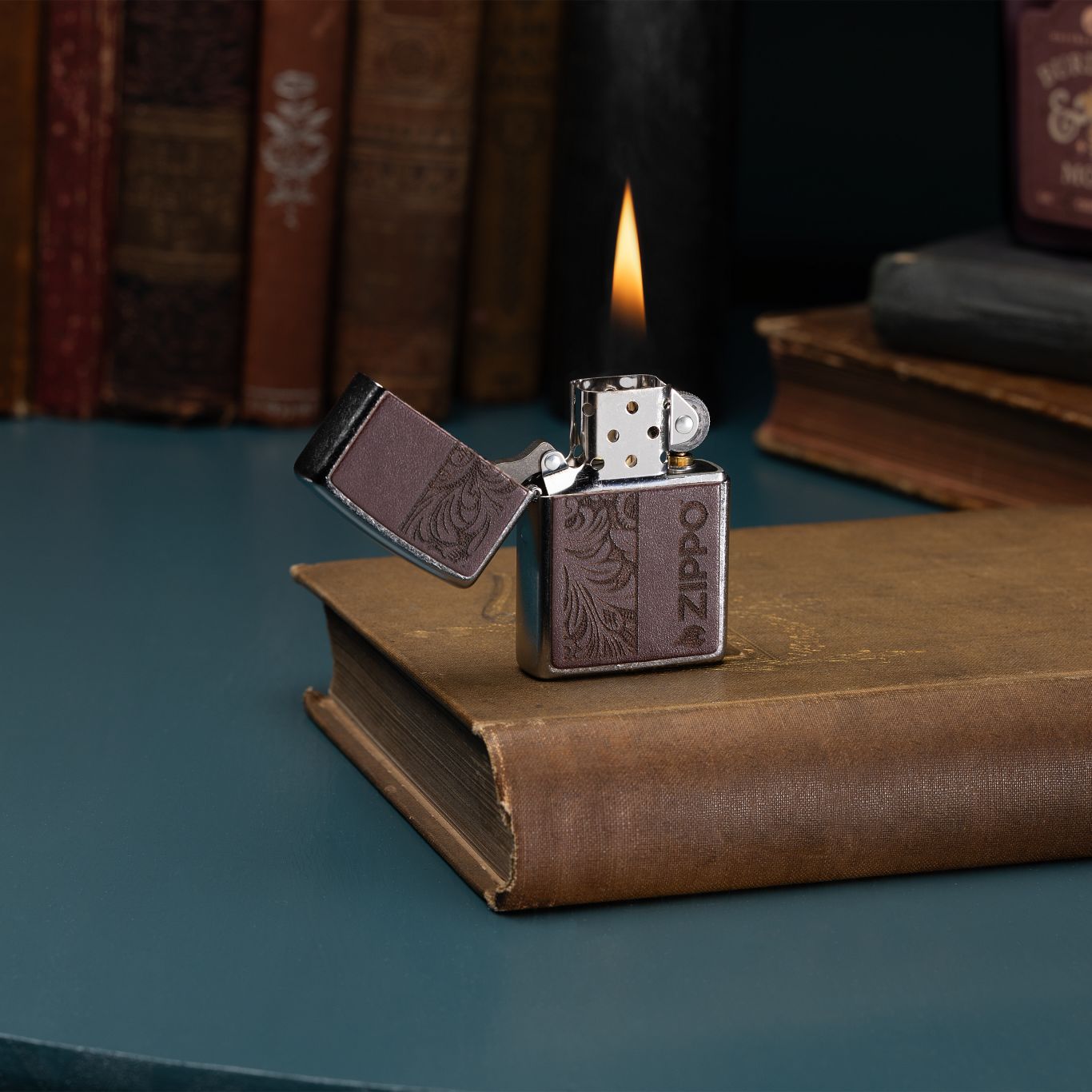 Lifestyle image of Zippo Brown Leather Emblem - Zippo Design Street Chrome Windproof Lighter with its lid open and lit standing on worn brown book with more books in the background.