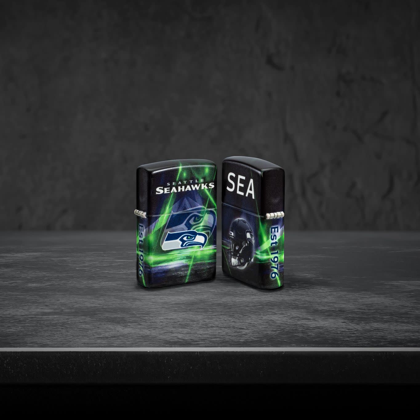 Lifestyle image of two Zippo NFL Seattle Seahawks 540 Matte Windproof Lighters, one showing the front of the lighter and the other showing the back, standing on a dark grey surface.