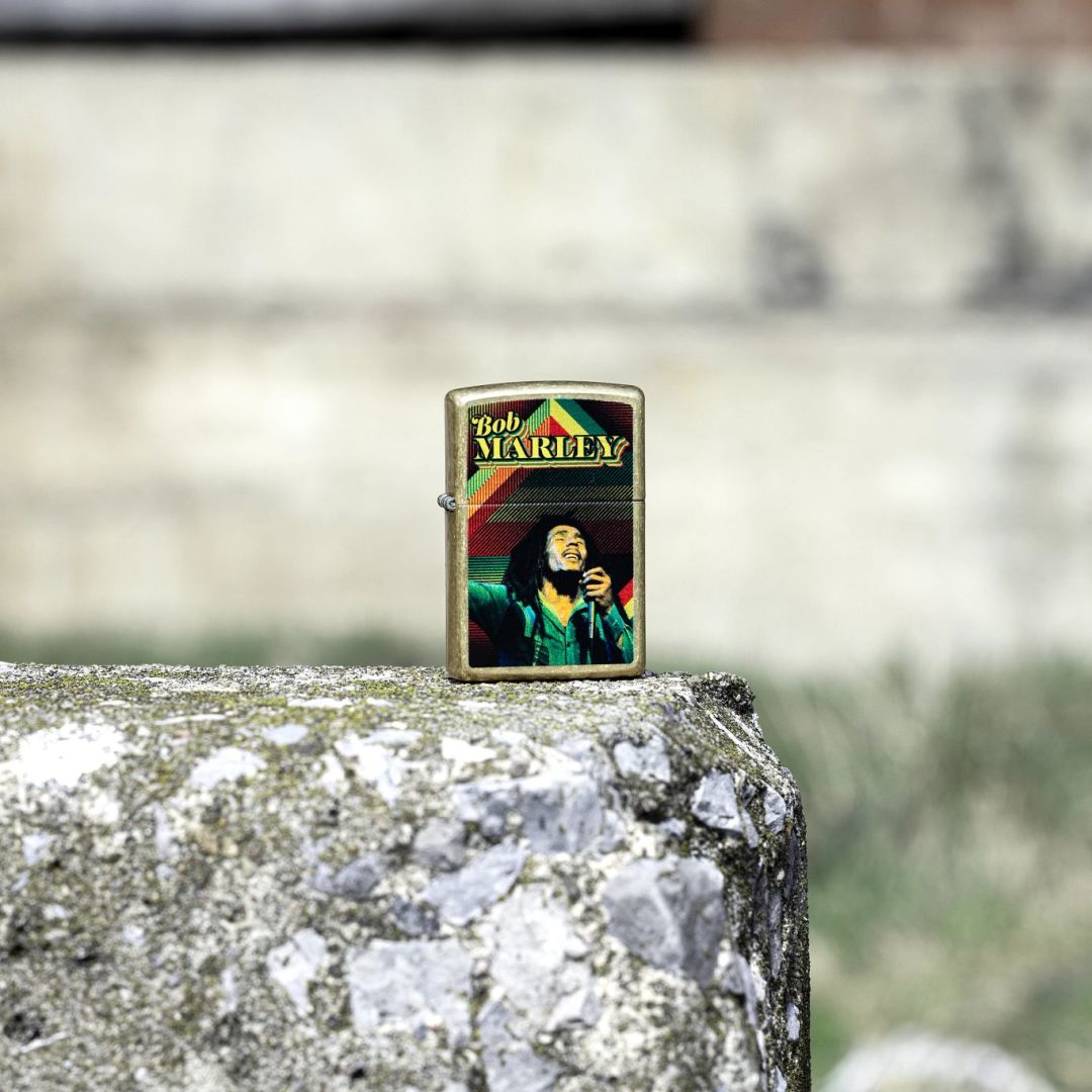 Lifestyle image of Zippo Bob Marley Soul Design Street Brass Windproof Lighter standing on a concrete surface showing the front of the lighter with an out of focus background. 

