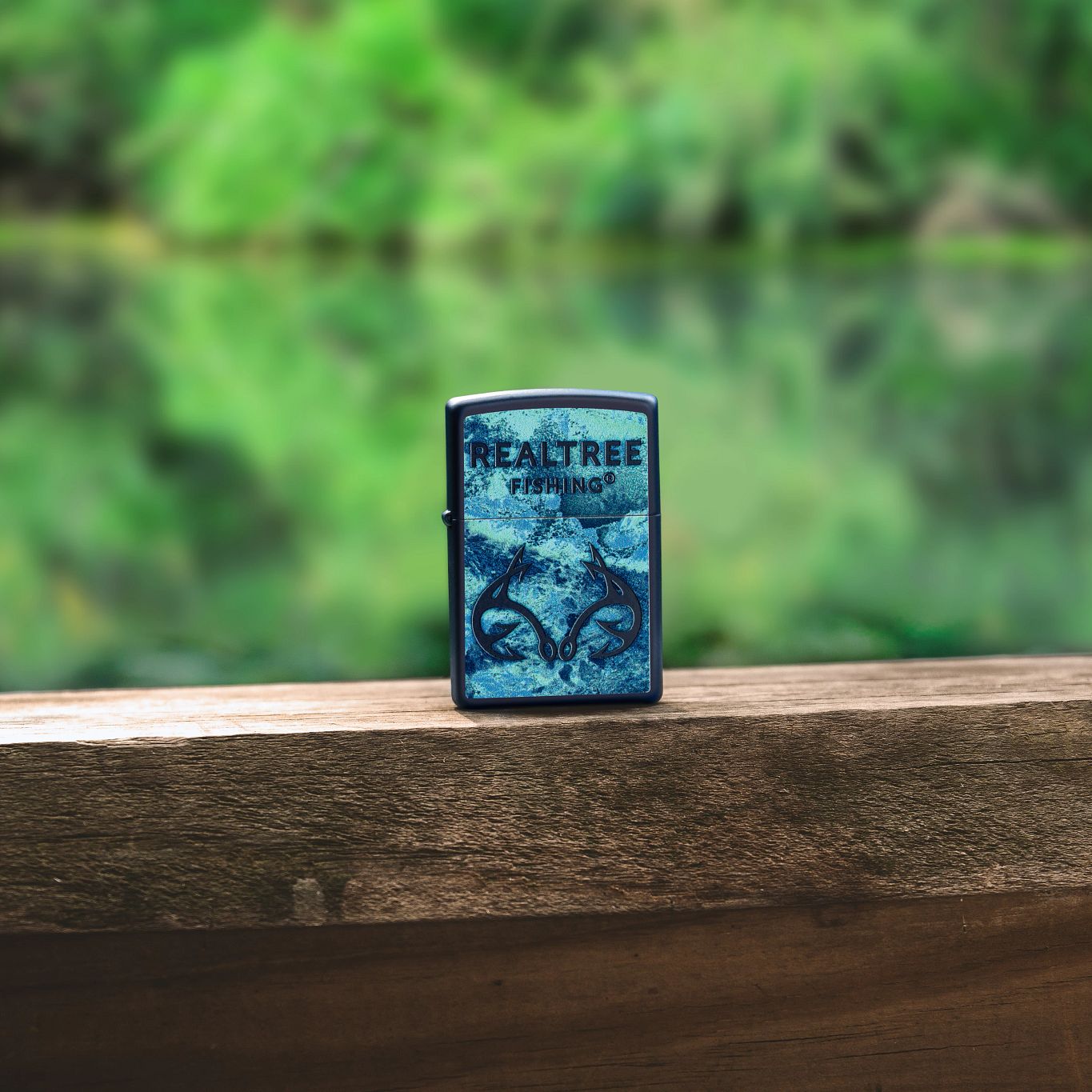Lifestyle image of Zippo Realtree® Fishing™ Design Navy Matte Windproof Lighter standing on a wooden railing with a blurred river and greenery background.

