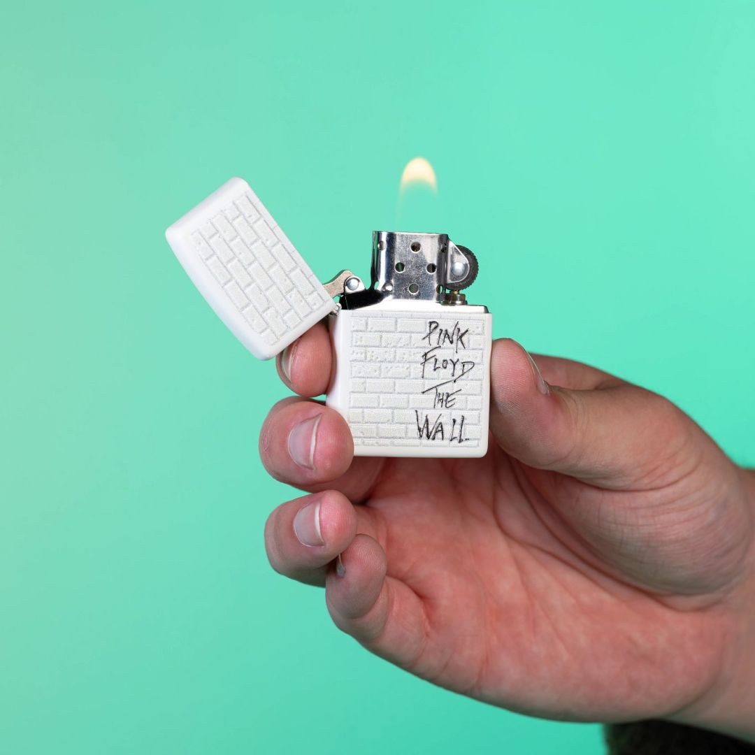 Lifestyle image of Zippo Pink Floyd The Wall Design White Matte Windproof Lighter lit in hand showing the front of the lighter with an aquamarine-colored background.

