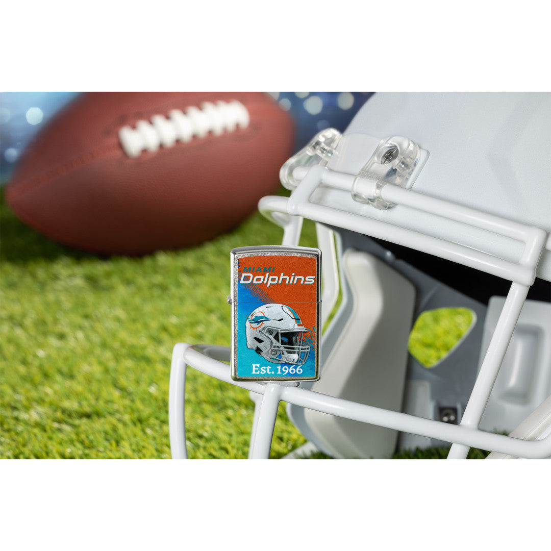 Lifestyle image of Zippo NFL Miami Dolphins Helmet Design Street Chrome Windproof Lighter standing on the facemask of a white helmet showing the front of the lighter with a football lying on a football field as the background.

