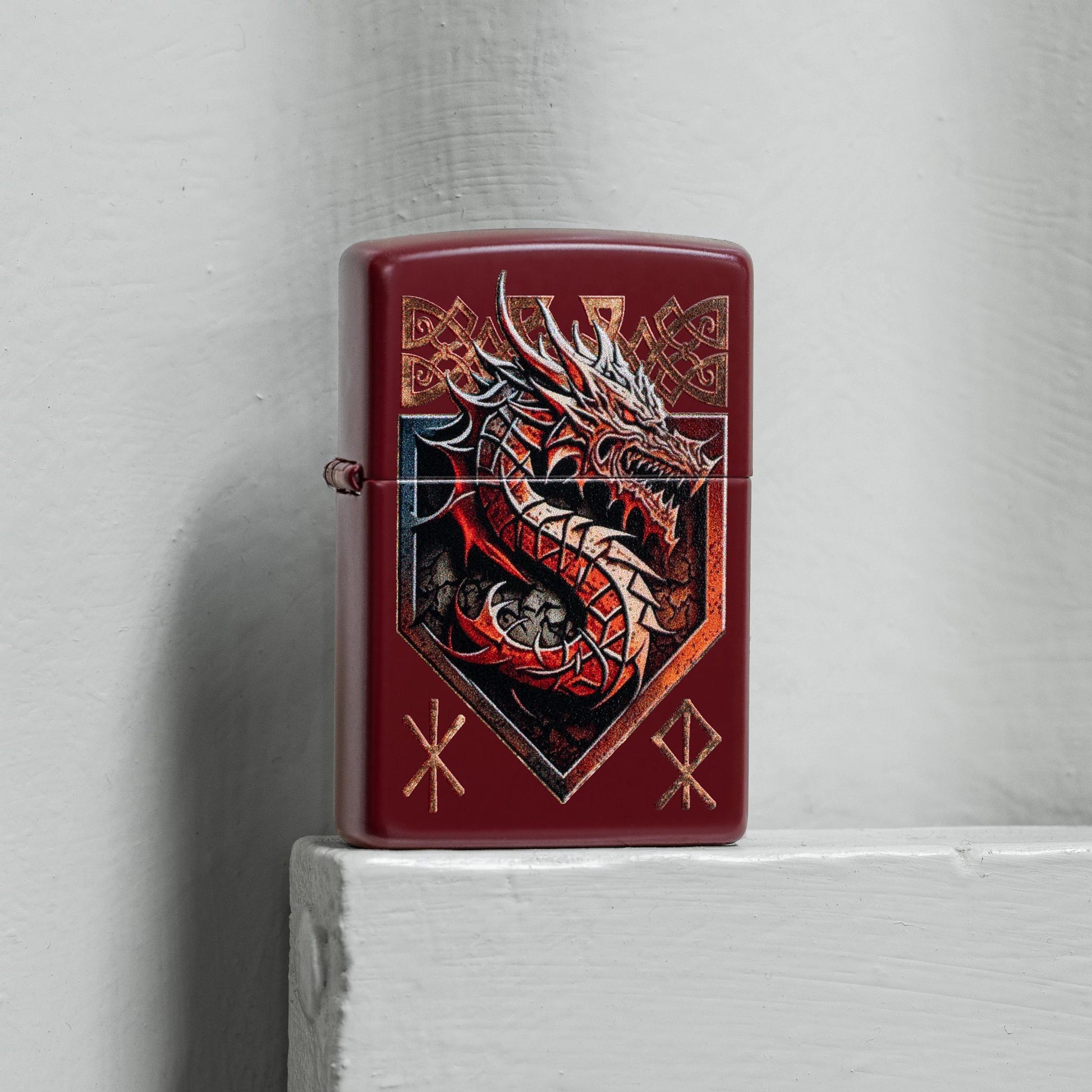 Lifestyle image of Zippo Dragon Shield Design Merlot Windproof Lighter standing on a light gray painted board next to a light gray painted concrete wall. 

