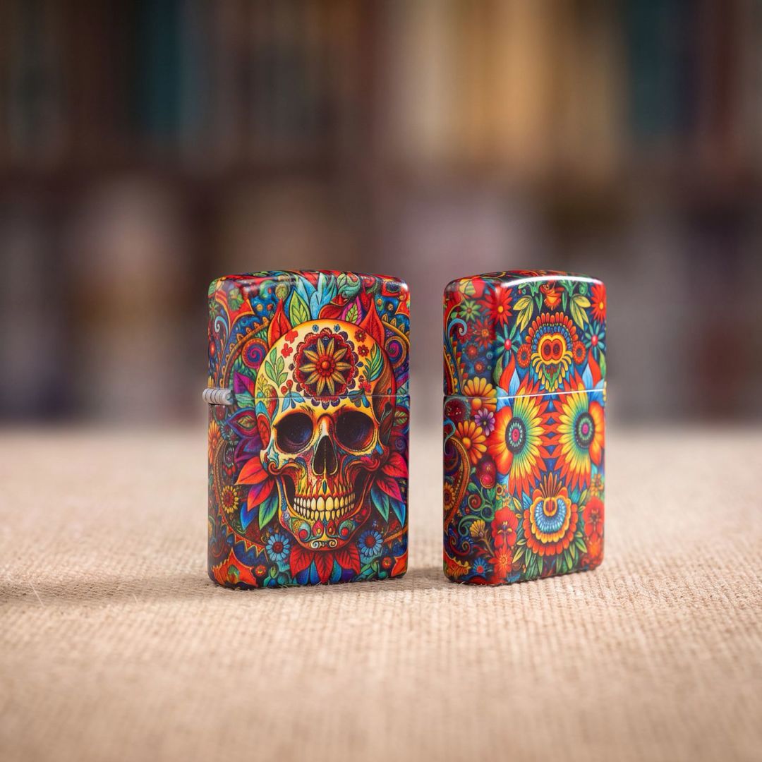 Lifestyle image of two Zippo Day of the Dead Skull Design 540 Matte Windproof Lighters standing on a tan placemat with an out of focus background. One showing the front and hinge side of the lighter while the other is showing the back and non-hinge side of the lighter. 


