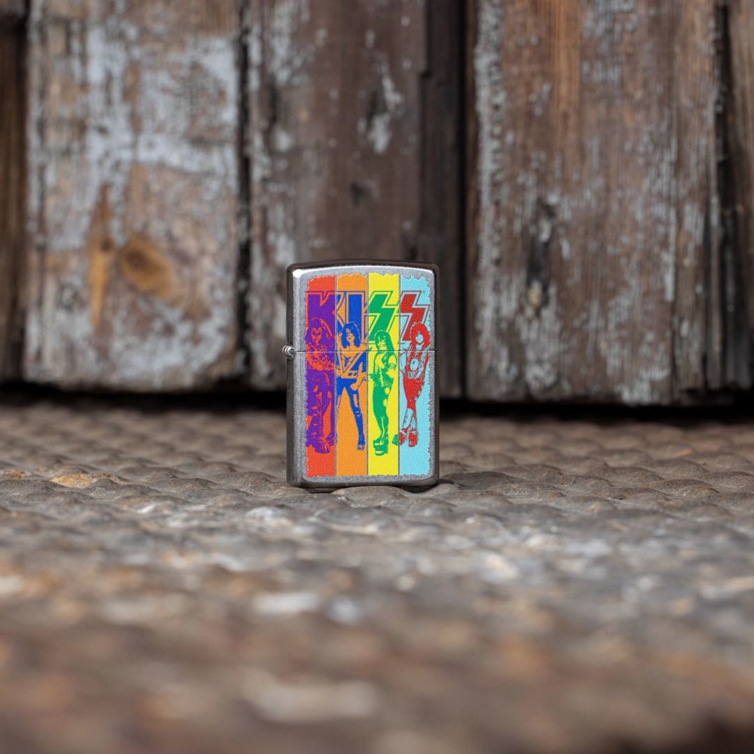 Lifestyle image of Zippo KISS® Fly Flag Design Street Chrome Windproof Lighter standing on a textured metal floor in front of an old, weathered wooden door showing the front of the lighter. 

