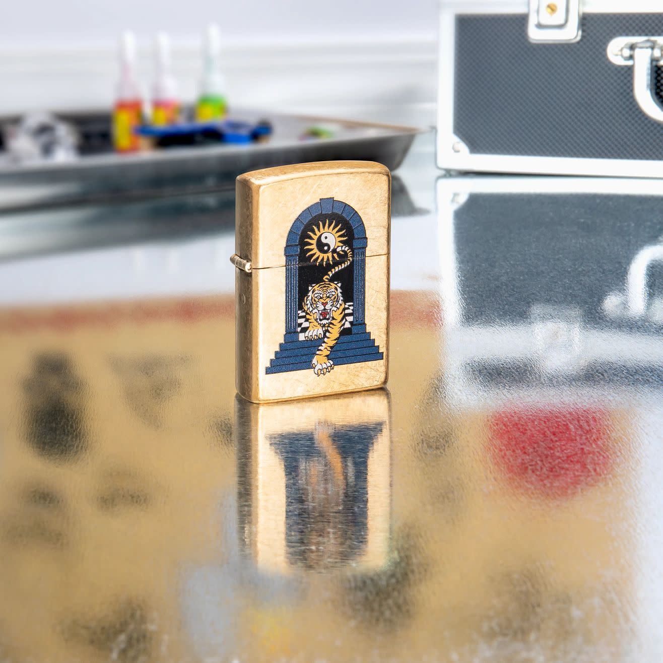 Lifestyle shot of Zippo Tiger Tattoo Design Tumbled Brass Windproof Lighter standing in on a reflective table with tattoo equipment.