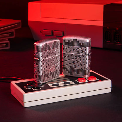 Lifestyle image of two Gaming Cheat Code ArmorÂ® Antique Silver Windproof Lighters standing on a vintage gaming controller with a retro gaming system in the background. One lighter is showing the front of the design with the other showing the back.