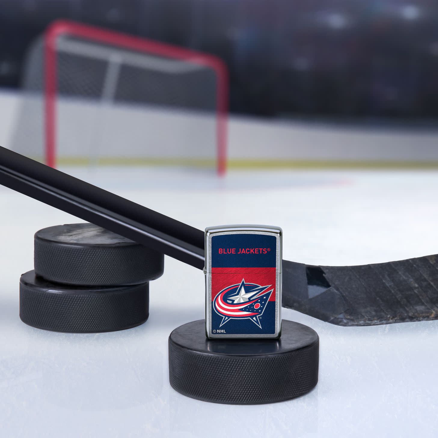 Lifestyle image of the NHL® Colorado Avalanche™ Street Chrome™ Windproof Lighter standing with a hockey puck and hockey stick, with a hockey net in the background.